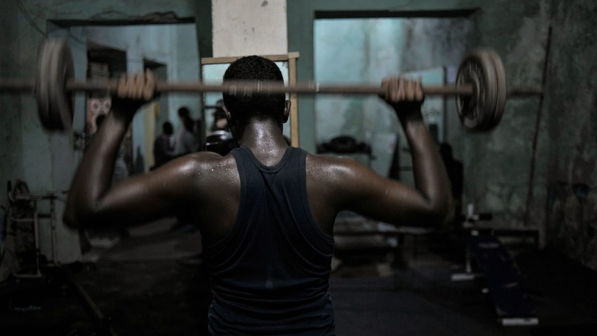 A man seen from behind lifting a barbell overhead in a dimly lit home gym.